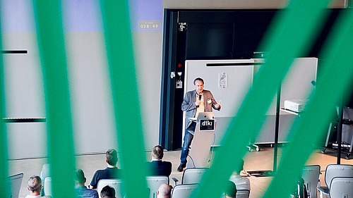 Das Bild zeigt einen Vortragssaal, in dem ein Mann an einem Rednerpult steht und vor Publikum spricht. Der Blick auf die Szene ist durch grüne vertikale Linien gerahmt, was eine besondere Perspektive erzeugt.