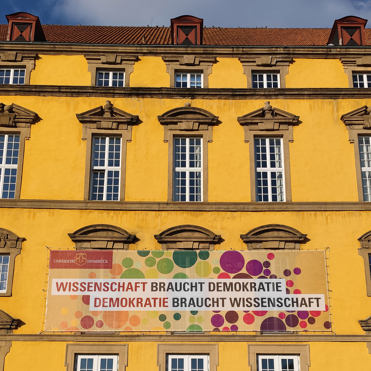 Blick auf ein großes buntes Banner am Osnabrücker Schloss. Aufschrift: Wissenschaft braucht Demokratie, Demokratie braucht Wissenschaft