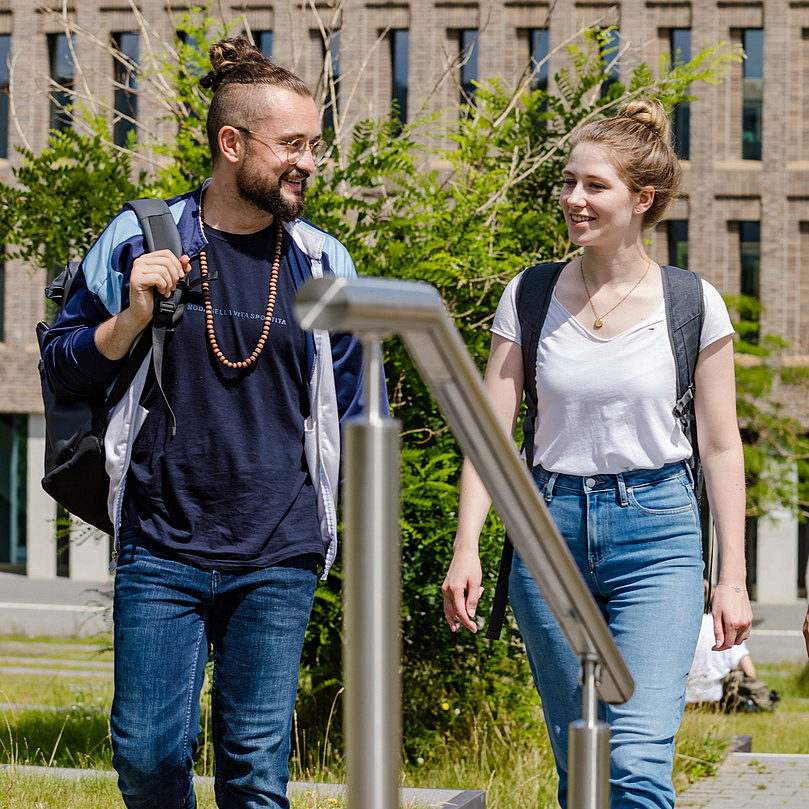 Vier Studierende unterhalten sich beim gehen auf dem Campus, im Hintergrund ist die Biliothek am Westerberg zu sehen