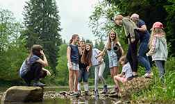 Von unten Blick auf lachende Jugendliche in lässigen Anzügen stehen auf Felsen im Fluss Wasser im Sommer Tag. Junge Freunde haben Spaß im Freien in der Nähe von grünen Wald. Konzept der Freundschaft, Natur, Camping.