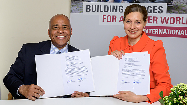 Prof. Dr. Susanne Menzel-Riedl and Dr. Roland S. Rochon pose for a photo in front of a stand with the title “Building Bridges for a better world. UOS international” while sitting for a photo. They are both holding a signed document in their hands and smiling at the camera.