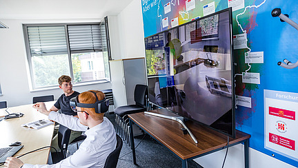 A man wears augmented reality glasses and reaches for something with his hand. Behind him, his view is transmitted to a screen. He is navigating a menu.