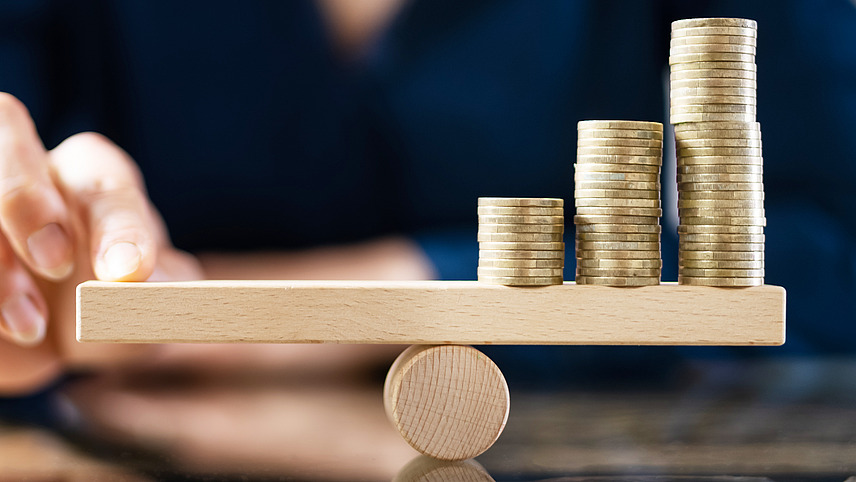 Several towers of coins stand on a wooden seesaw on the right, while a finger keeps the balance on the left.