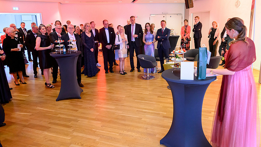 Numerous guests stand together and listen to a speech by Susanne Menzel, who is standing at a bar table in the foreground