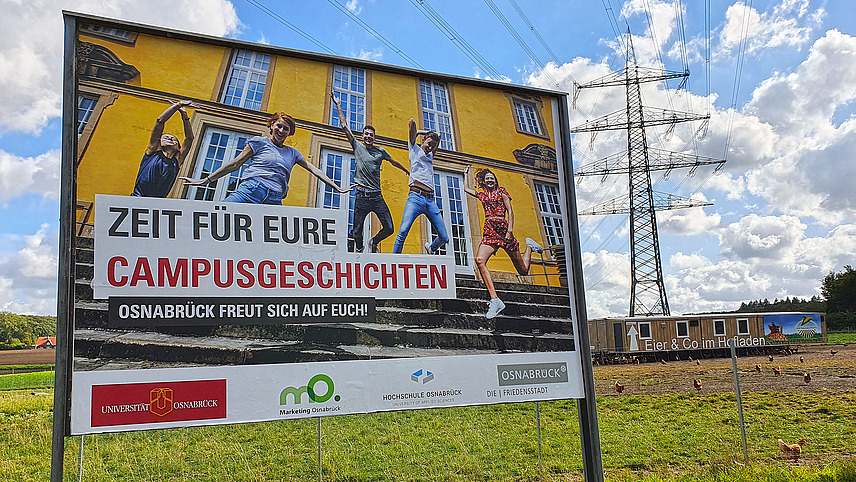 A large advertising poster showing students jumping into the air on the steps of the castle. The inscription reads “Time for your campus stories. Osnabrück is looking forward to seeing you!”.