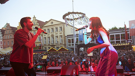 Two people stand opposite each other on stage and look at each other while singing