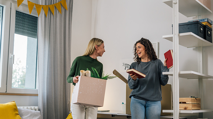 Zwei Studentinnen stehen in einem Zimmer und räumen ein Bücherregal ein.