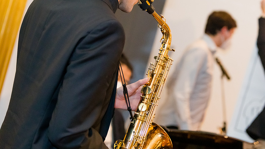 An elegantly dressed man plays the saxophone in front. The outlines of the rest of the orchestra can be seen in the background. All the people are elegantly dressed and, with the exception of the saxophone player, are wearing medical masks.