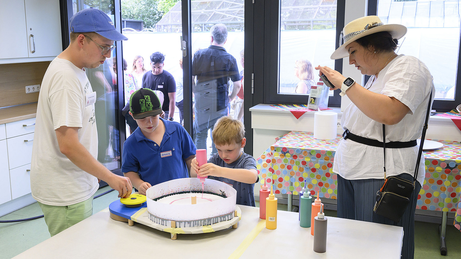 Two adults and two children are standing at a table, the children are crafting with paint bottles.