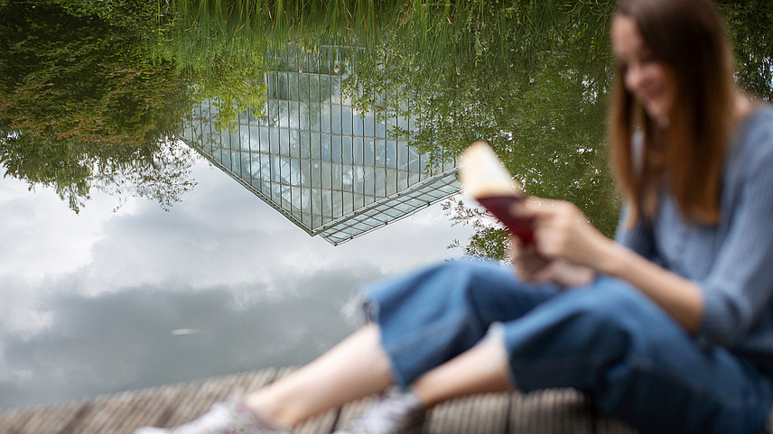 Eine Frau liest ein Buch auf einer Holzplatform vor einem Teich. In der Reflektion des Wassers ist die Spitze des Regenwaldhauses zu erkennnen.