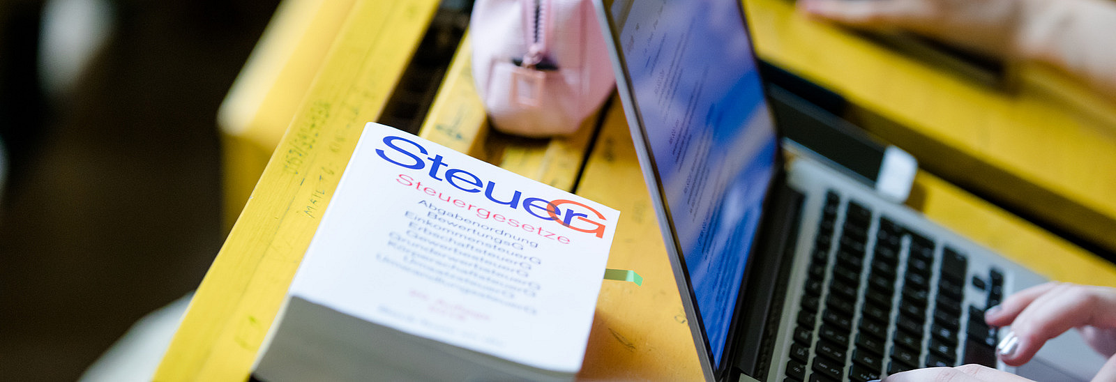 Blick auf einen Klapptisch in einem Hörsaal. Eine Studentin tippt an ihrem Laptop. Daneben liegt ein Buch mit dem Titel: "Steuergesetze".