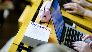 Blick auf einen Klapptisch in einem Hörsaal. Eine Studentin tippt an ihrem Laptop. Daneben liegt ein Buch mit dem Titel: "Steuergesetze".