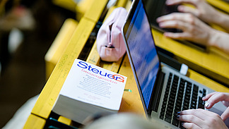 Blick auf einen Klapptisch in einem Hörsaal. Eine Studentin tippt an ihrem Laptop. Daneben liegt ein Buch mit dem Titel: "Steuergesetze".