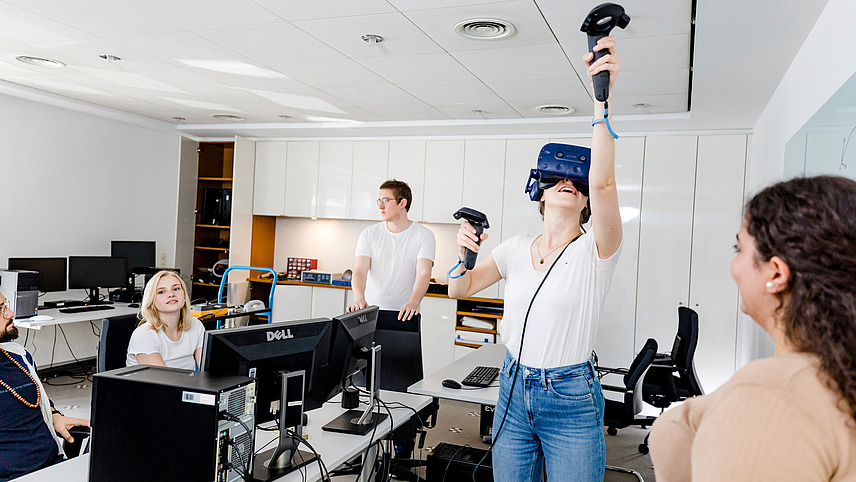 A student is wearing a VR headset and the corresponding controllers in her hands, she is trying to reach something high up. A few students are standing and sitting around her.