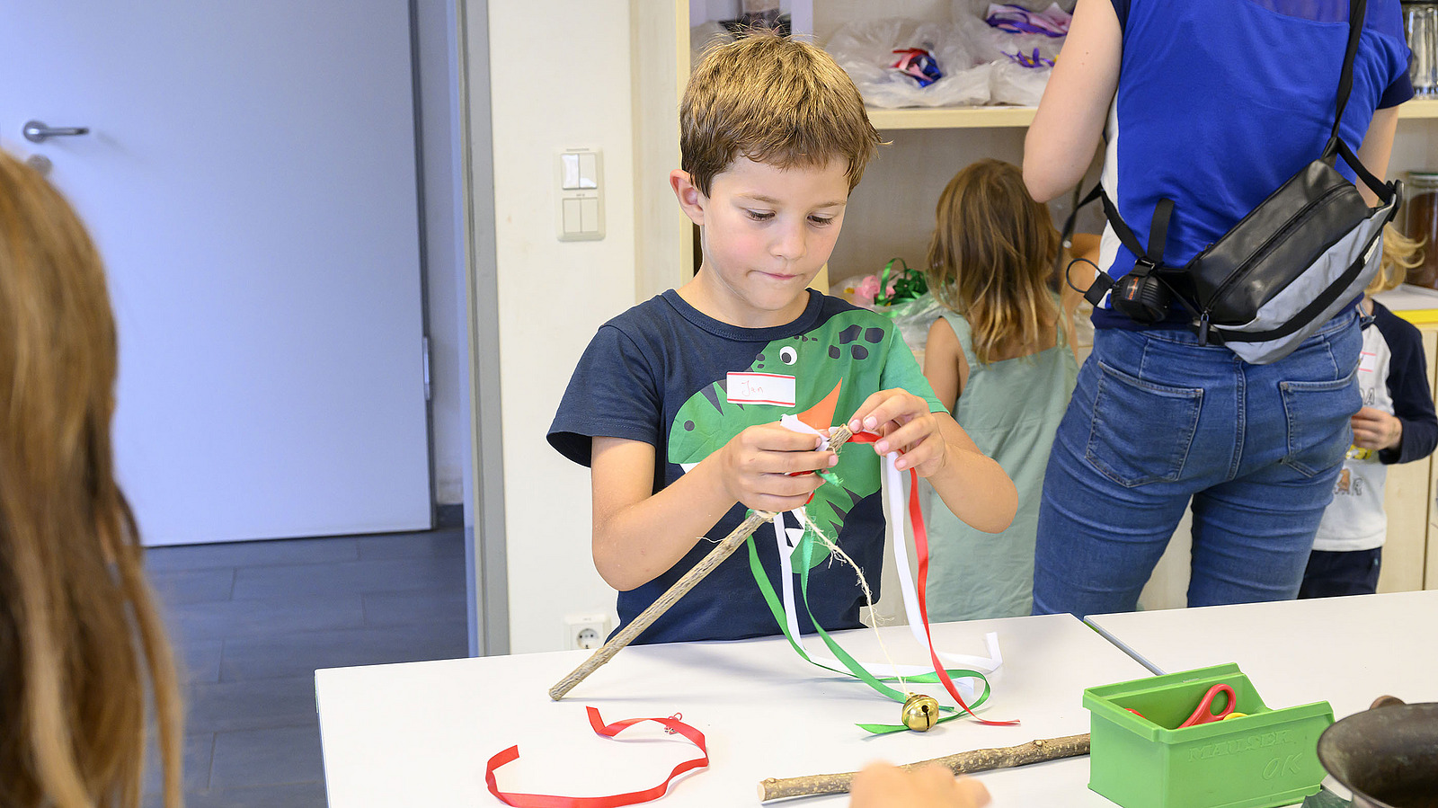 A boy crafts at a table with ribbons and sticks.