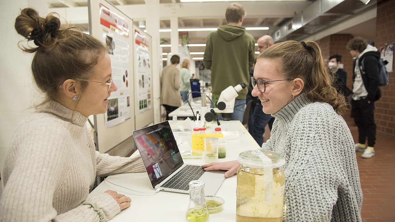 Two women are sitting at a table with a laptop in front of them. The woman on the left is wearing a light sweater and has her hair in a ponytail, while the woman on the right is wearing a gray knitted sweater. Both are smiling and appear to be chatting. On the table, there are various jars with green samples and a microscope in the background. In the background, there are additional people and posters visible.