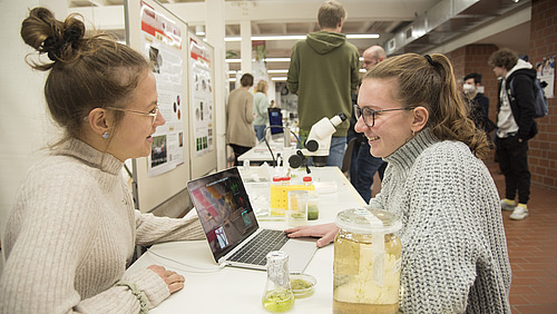 Two women are sitting at a table with a laptop in front of them. The woman on the left is wearing a light sweater and has her hair in a ponytail, while the woman on the right is wearing a gray knitted sweater. Both are smiling and appear to be chatting. On the table, there are various jars with green samples and a microscope in the background. In the background, there are additional people and posters visible.