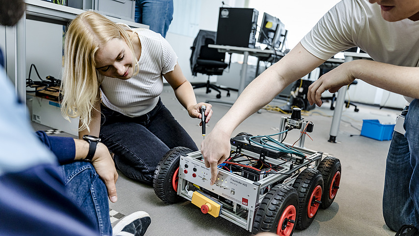 A few students are kneeling in front of a box on wheels in a workroom. Many cables are sticking out of the box and there are many buttons and switches on one side. A student operates one of the switches.