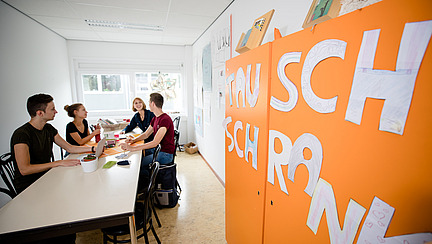 In a bright room, several people are sitting at a long table chatting. On a cupboard in the foreground hangs an orange poster with the slogan “Exchange cupboard”. 