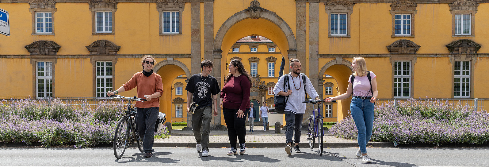 Fünf internationale Studierende in sommerlicher Kleidung überqueren eine Straße vorm Schlossgebäude der Universität Osnabrück. Zwei Personen schieben dabei ihre Fahrräder.