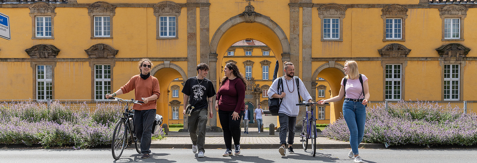 Five international students in summer clothing cross a street in front of the Osnabrück University castle building. Two people are walking with their bikes.