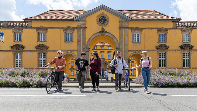 Fünf internationale Studierende in sommerlicher Kleidung überqueren eine Straße vorm Schlossgebäude der Universität Osnabrück. Zwei Personen schieben dabei ihre Fahrräder.