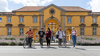 Five international students in summer clothing cross a street in front of the Osnabrück University castle building. Two people are walking with their bikes.