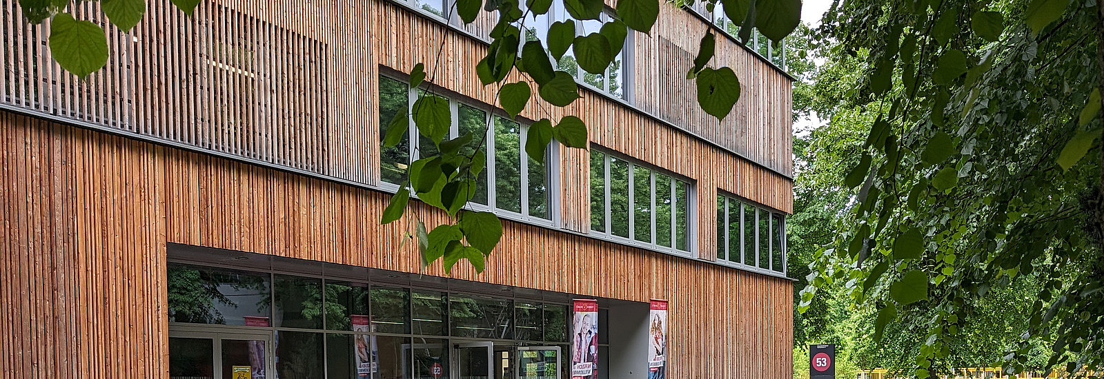 Ein modernes Gebäude mit einer Fassade aus Holzlatten und großen Fenstern ist im Bild zu sehen. Vor dem Eingang sind zahlreiche Fahrräder abgestellt. Die Umgebung wird von Bäumen und Grünpflanzen gesäumt, die dem Bereich eine natürliche Atmosphäre verleihen. Auf einem Schild neben dem Eingang ist die Nummer 53 zu erkennen.