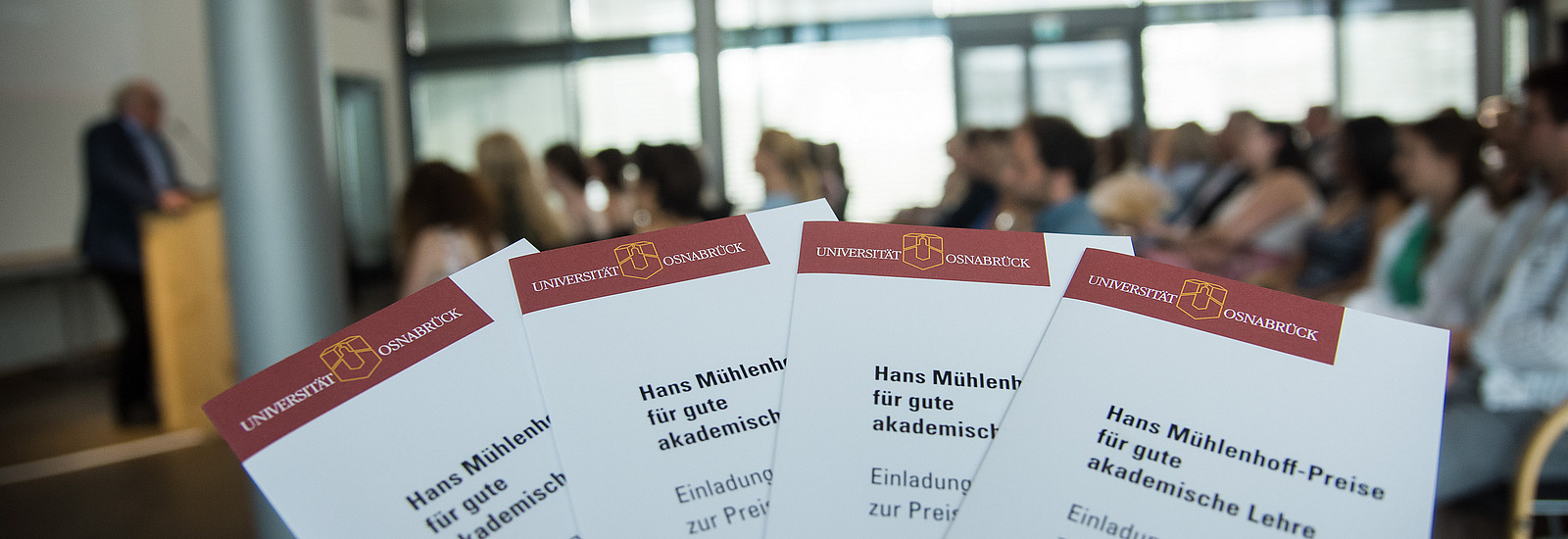 Four flyers for the Mühlenhoff Prize for Good Academic Teaching are in the foreground, behind them a hall with audience and speakers.