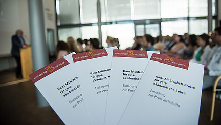 Four flyers for the Mühlenhoff Prize for Good Academic Teaching are in the foreground, behind them a hall with audience and speakers.