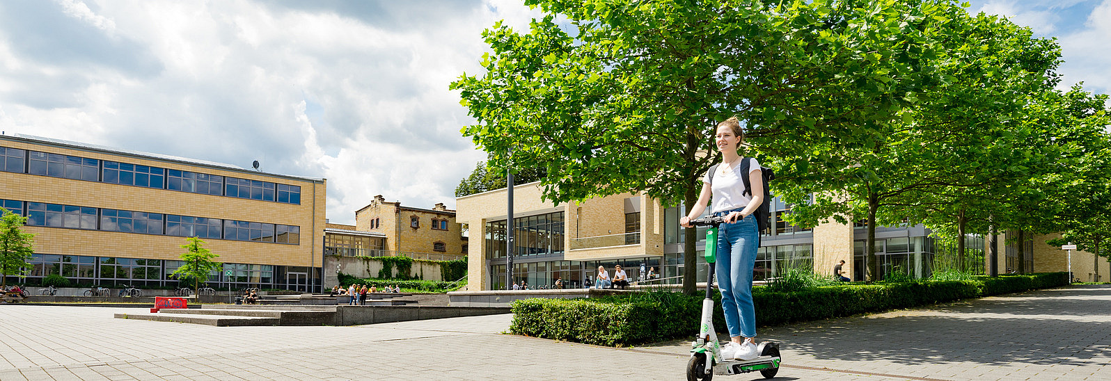 A young woman rides an electric scooter across a paved square surrounded by modern buildings and trees. Various people can be seen sitting or moving around in the background, while the sky is partly cloudy.