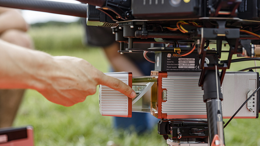 Close-up of a drone. A person points with their finger to a part of the lower extension, which also contains the camera. A meadow and a field can be seen in the background.