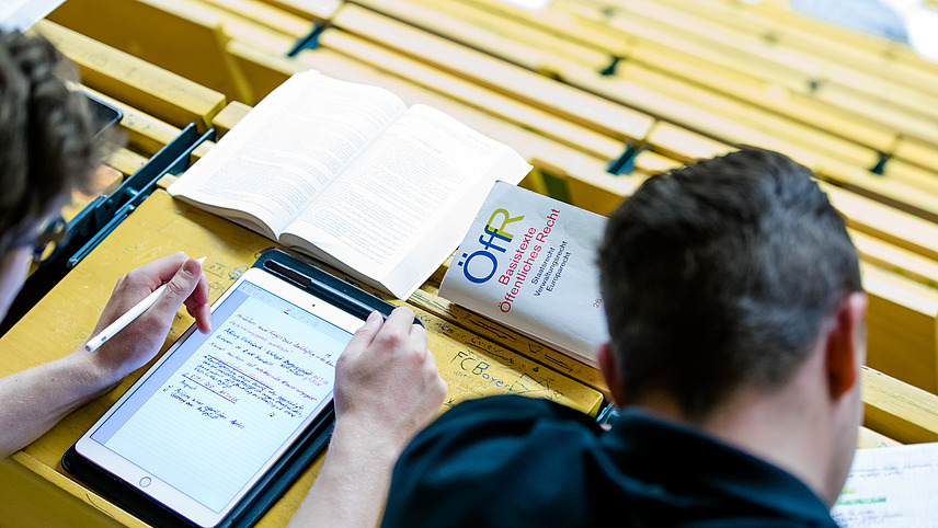 Zwei männliche Studenten sitzen in einem Hörsaal und machen sich Notizen auf einem Tablet und Papier. Vor ihnen liegen mehrere Bücher, eines mit dem Titel „Grundlagentexte des öffentlichen Rechts. Verfassungsrecht, Verwaltung und Europarecht“.