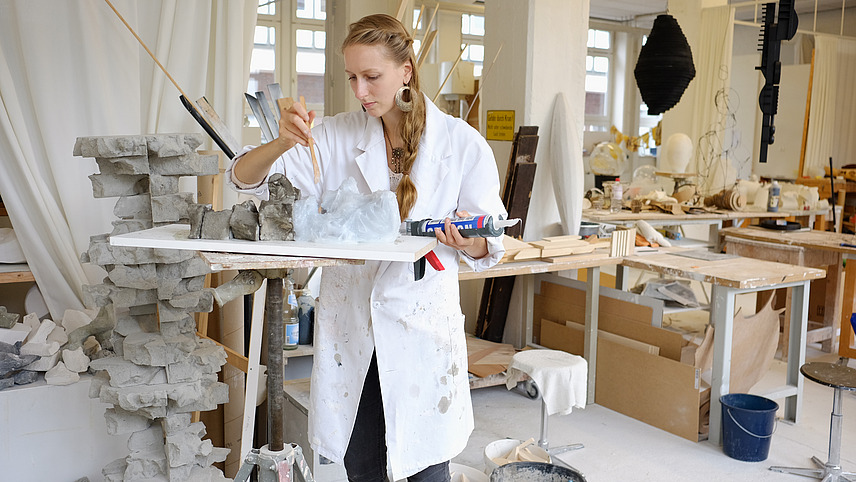 A blonde woman in a long white coat is working in an art and craft studio on a sculpture that resembles stacked stone blocks. She is working with a glue. Next to her is a bucket in which the material for the sculpture is being mixed.