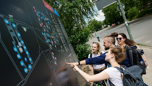 Ein paar Studierende zeigen mit dem Finger auf Gebäude auf einem großen Lageplan vom Westerberg