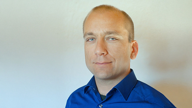 A man with short, blond hair and a blue shirt looks directly into the camera. His facial expression is neutral and he has a slight smile.