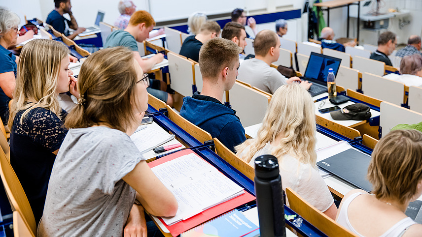 Students of different ages sit in a lecture hall and take notes or work on their laptops while listening to the lecture.