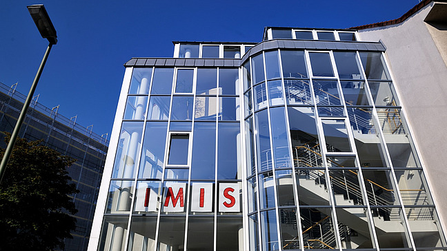 The picture shows the façade of a modern building with large glass windows. Above the windows is the lettering “IMIS” in red letters. The building has an interesting architecture, with a curved part and visible stairs within the transparent structure.