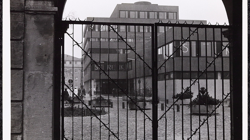 View of a building through a wrought-iron gate. Posters with the individual letters of the word STREIK are displayed in the windows