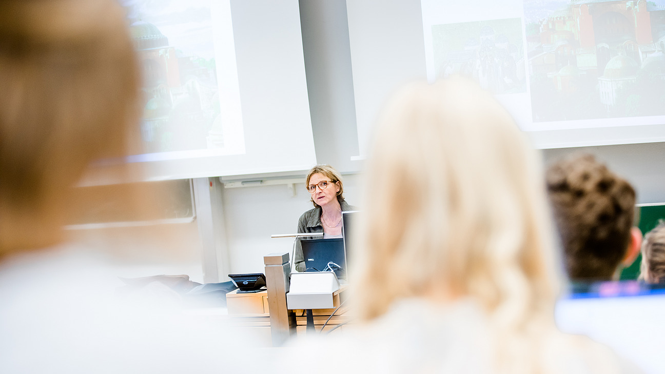 A lecturer sits at a table and speaks to a group of students, some of whom can be seen out of focus in the foreground. 