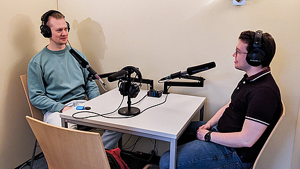 Two men are sitting at a table with microphones, wearing headphones and talking.