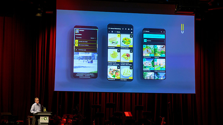 Eine Person steht an einem Rednerpult auf der Bühne, über ihm auf der Leinwand sind drei Smartphones mit der App des Studierendenwerks zu sehen