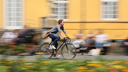 Ein junger Mann fährt auf einem schwarzen Fahrrad. Die Bewegung des Fahrrads wird durch eine Verwischung im Hintergrund deutlich, die Menschen und eine gelbe Fassade zeigt. Bunte Blumen sind im Vordergrund zu sehen.