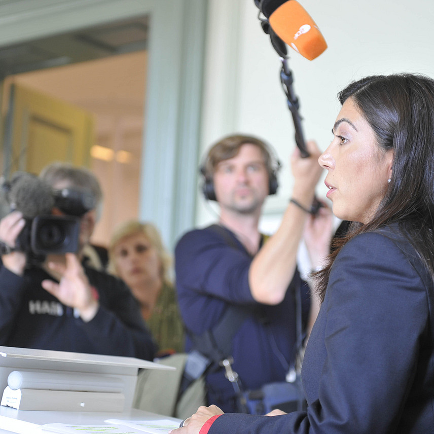 Eine Frau in formeller Kleidung spricht in ein Mikrofon während einer Pressekonferenz. Im Hintergrund sind mehrere Journalisten und Kameraleute zu sehen, wobei eine Person Videoaufnahmen macht. 