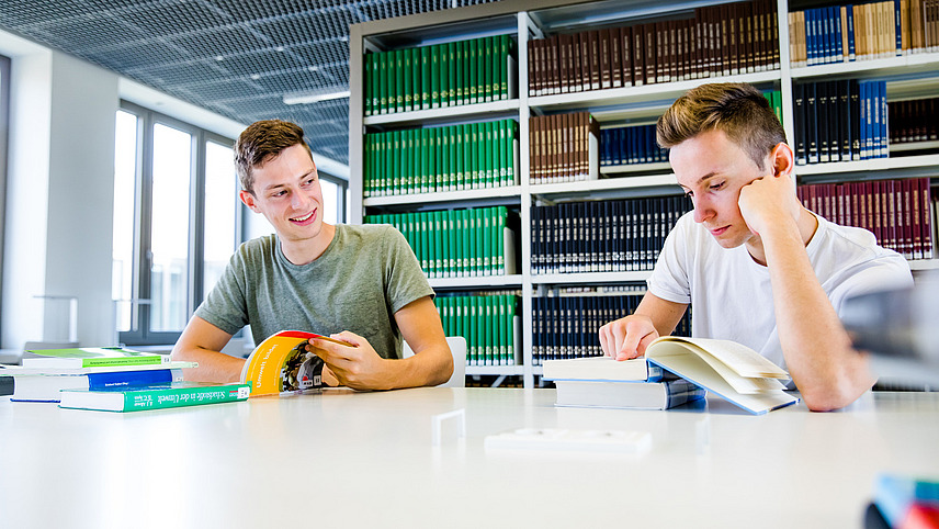 Zwei Studenten sitzen an einem Tisch in der Bibliothek am Westerberg und unterhalten sich. Vor ihnen liegen Bücher.