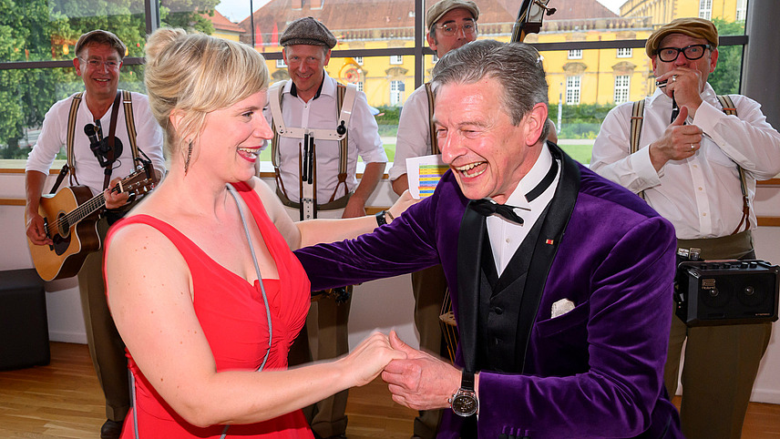 Presenters Nele Kießling and Michael Hull stand laughing and dancing in front of the marching band