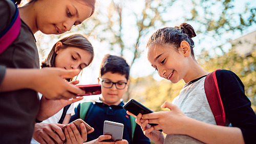 Kids playing video games on smart phone after school Schülerinnen und Schüler stehen auf dem Pausenhof und blicken auf ihre Smartphones.