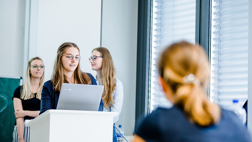 Drei Studentinnen stehen in einem Seminarraum vor einem Podium. Eine von ihnen schaut auf einen Laptop und spricht, während das Publikum zuhört.