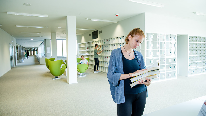 Eine Studierende steht in bequemer Kleidung in einer Bibliothek und liest in einem Buch. Im Hintergrund sucht ein Studierender sich ein Magazin aus einem Magazinständer während ein anderer Studierender in einem Sessel liest.