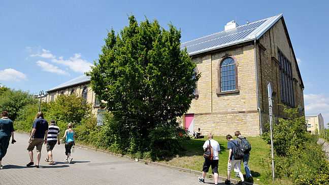 The picture shows several people walking towards a historic building. The building has a stone facade and large windows. There is a large green shrub in front of the building